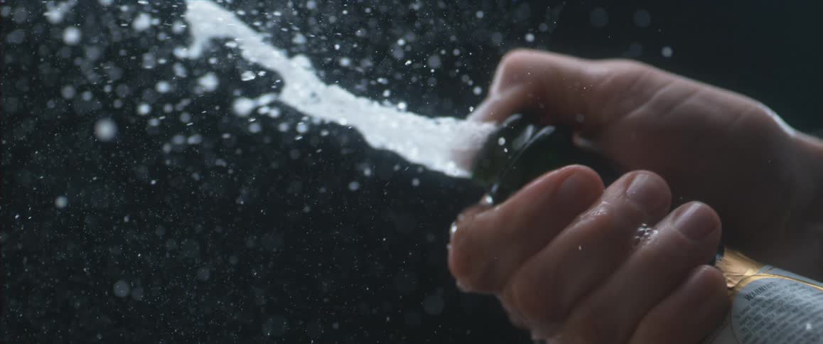 A person holds their thumb over a bottle while the carbonation fizzes out in slow motion