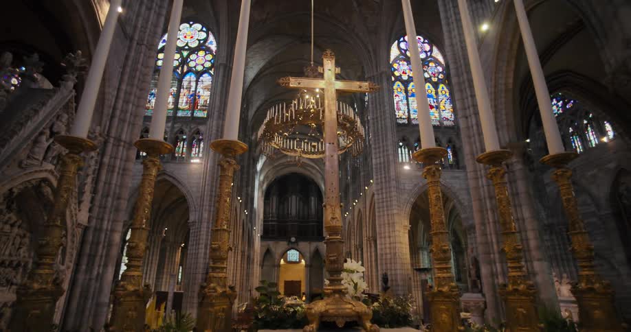 A slow pan across the interior of a large cathedral shows an altar with a large wooden cross and massive gold candle holders in the center with towering vaulted ceilings and colorful stainedglass windows in the background