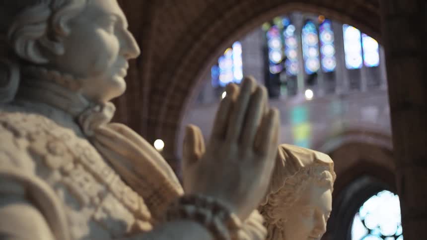 The video shows a closeup of two marble statues in a cathedral with stained glass windows in the background the camera pans slowly from the face of a female statue to a male statue wearing a crown both with their hands clasped in prayer