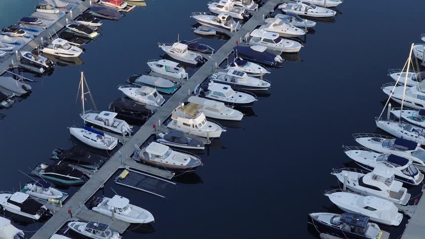 The video shows an aerial view of a marina with numerous boats and yachts of various sizes docked in neat rows the water is a deep dark blue reflecting the light and creating a sense of calm