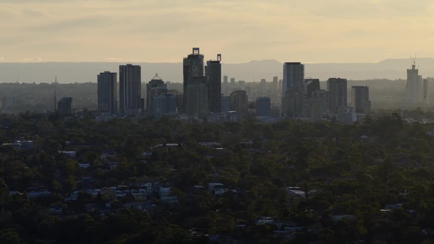 The video shows an establishing shot of a modern city skyline at dusk with numerous skyscrapers rising above a forest of trees the hazy golden light of the setting sun bathes the scene
