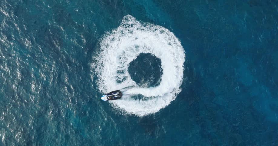 A person wearing a black diving suit rides a jet ski turning in a circle before speeding off into the ocean at midday