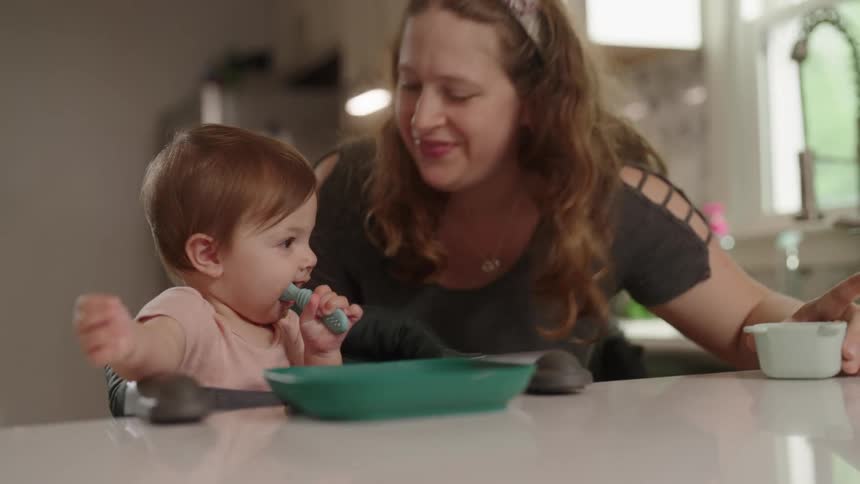A mother plays with her toddler son who is holding a spoon and eating a meal on the kitchen countertop at midday