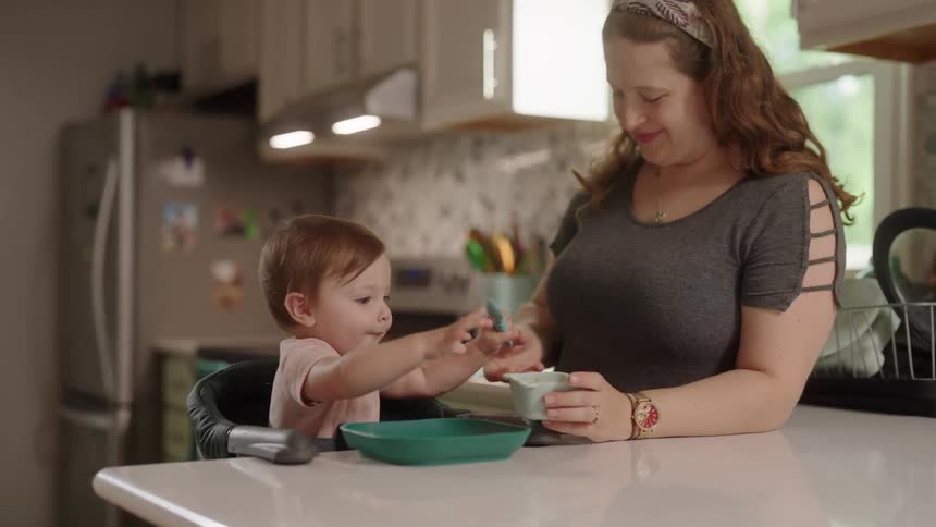 A mother stands beside her toddler son who is playing with his meal on the kitchen countertop at midday