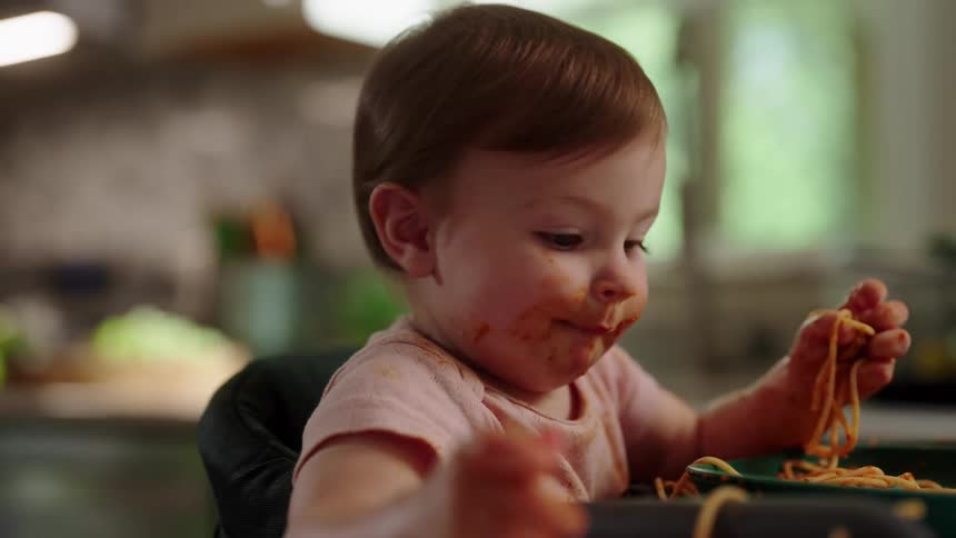 A toddler eats spaghetti with sauce on their face