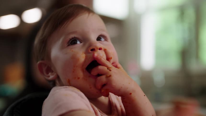 A baby with sauce on their face reaches for spaghetti while seated in a high chair