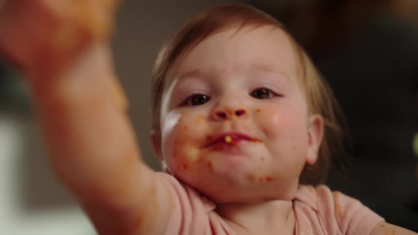 Toddler eats messily in a highchair while the mother adds more food to the dish