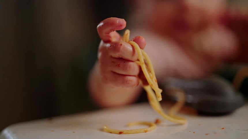 A toddlers small hand grasps strands of spaghetti coated in sauce