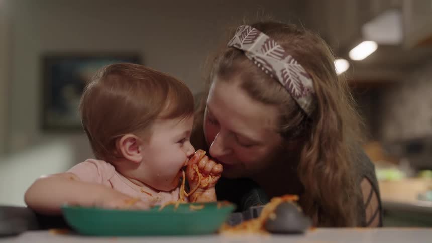 Mother kissing her baby who is eating pasta with sauce with her hands