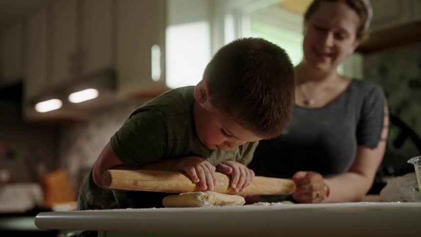 A young boy leans over to press a rolling pin into dough as a smiling woman watches