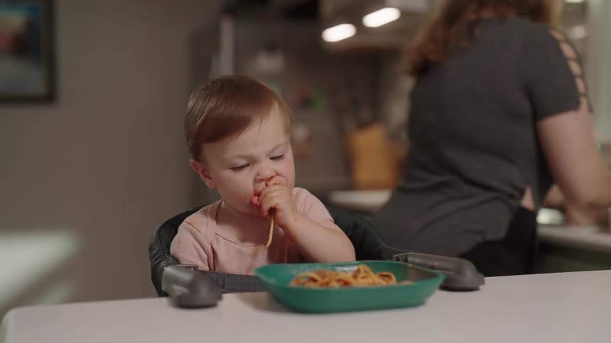 A small child in a high chair is messily eating a bowl of spaghetti with their hands in the background a woman is seen working in the kitchen