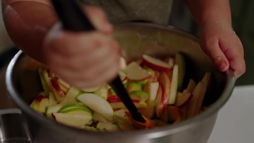 A childs hands are shown from above holding a large stainless steel bowl and using a plastic spatula to stir together a mix of sliced red and green apples