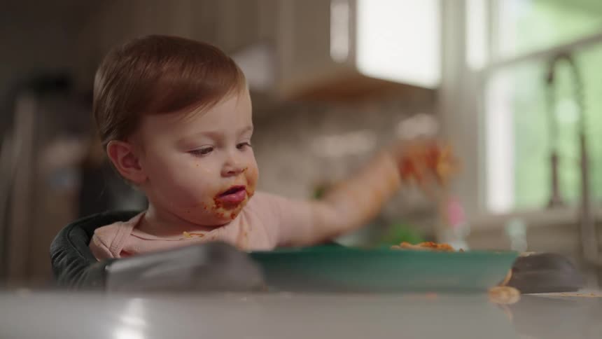 A toddler plays with a spaghetti meal on a plate in front of him on the kitchen countertop while the mother is busy with chores behind him at midday