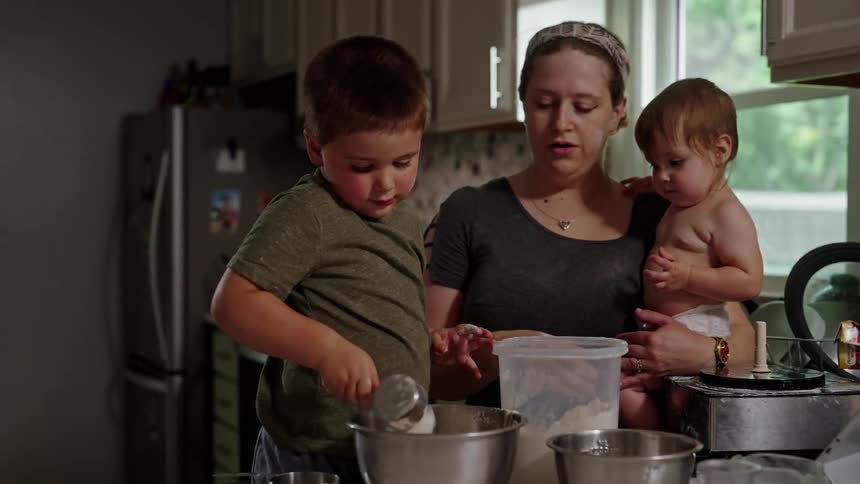 A woman holds a baby while watching a young boy scoop flour into a bowl in the kitchen