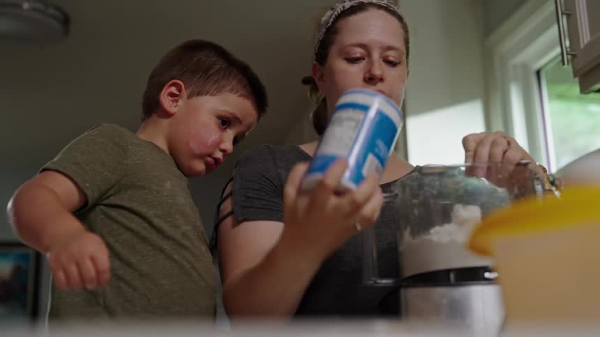 A woman measures ingredients into a food processor while a young boy watches and points