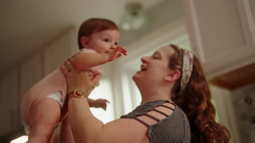 A woman picks up her baby and gently tosses him her in the air making her smile while in the kitchen at midday