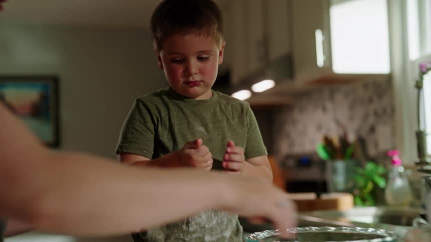 A boy helps his mother in the kitchen by cleaning a bowl with his palm while she makes dough on the countertop