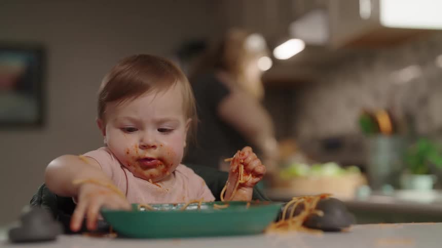 A toddler eating and playing with a spaghetti meal from a plate on the kitchen countertop while the mother prepares a meal behind her at midday