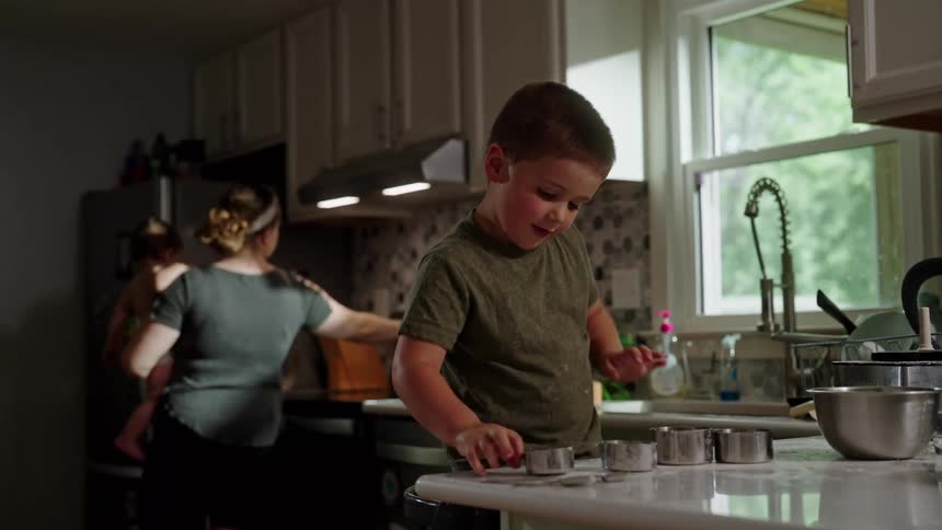 Boy playing in the kitchen while his mother is in the background with a baby on her lap