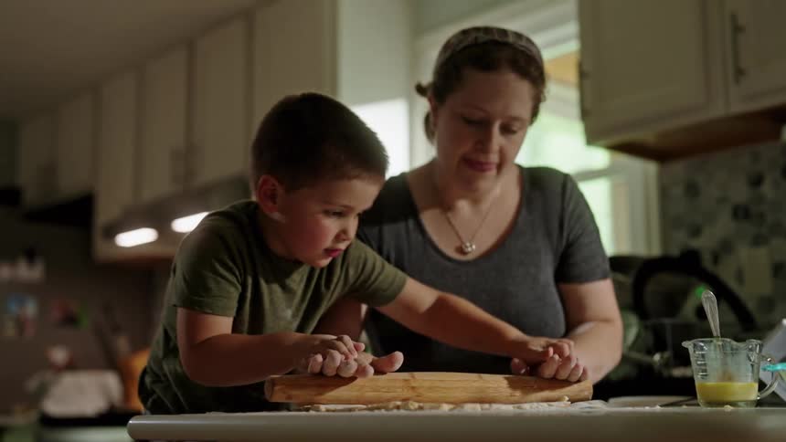 A woman guides a young boy as he rolls out dough with a rolling pin in the kitchen