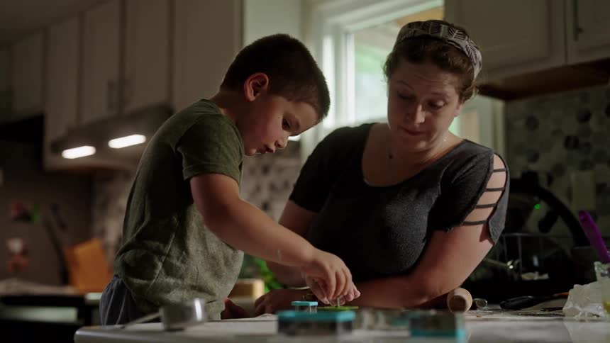 A young boy uses a cookie cutter while a woman watches and assists at the kitchen counter