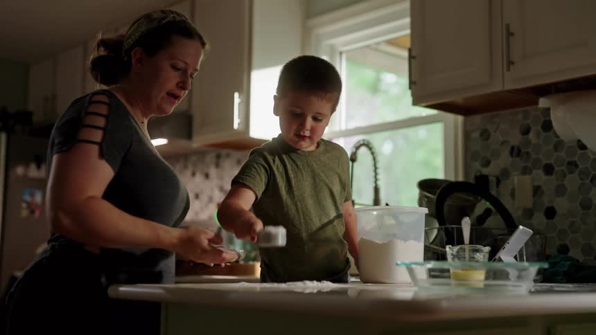 A mother has her son help her in the kitchen as he plays with flour inside a spoon the mother takes the flour pours it into a container and cleans the countertop of the excess flour