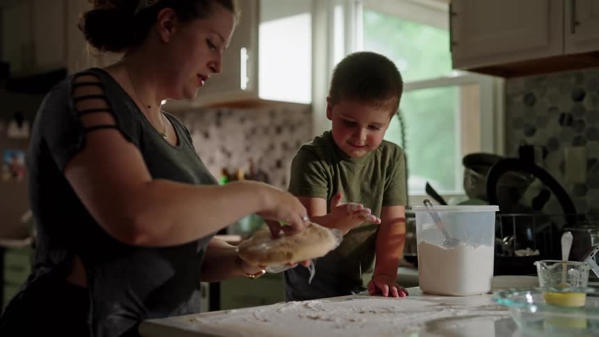 A woman and a young boy smile while preparing dough together in a warmly lit kitchen