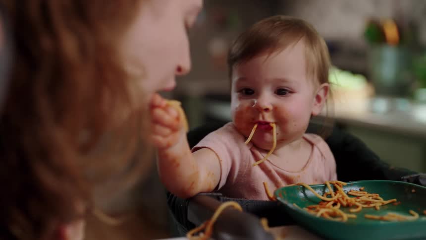 Baby in a high chair eating a bowl of spaghetti the child is covered in sauce and noodles making a mess as they eat while mother leans in to nibble