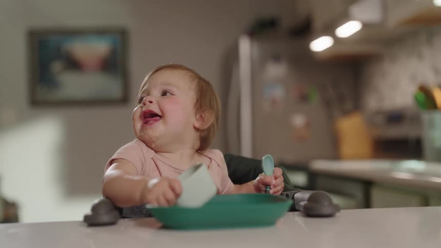 A smiling toddler sits at the counter holding utensils with a bowl in front of them