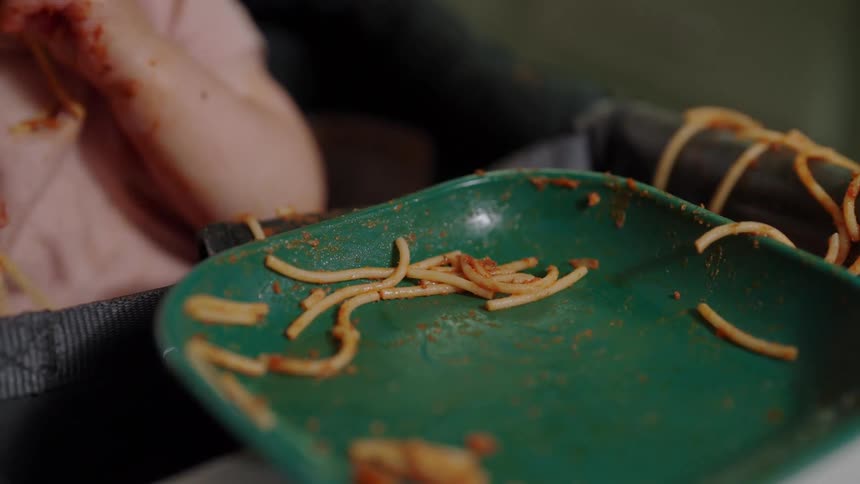 A closeup shows a babys hand covered in red sauce as they play with spaghetti in a green plate on a high chair tray