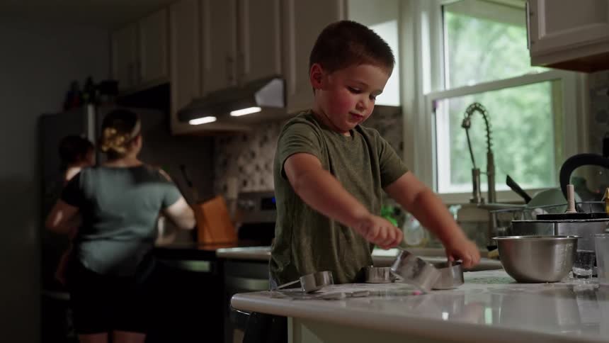 A kid playing with kitchen utensils on the countertop while the mother prepares a meal behind him at midday