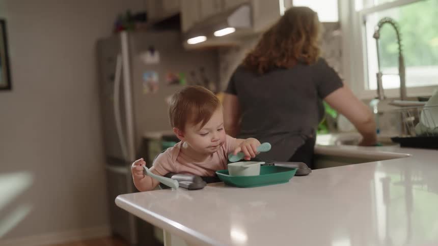 A mother is washing dishes at the kitchen sink while a child sits behind the countertop eating a meal at midday