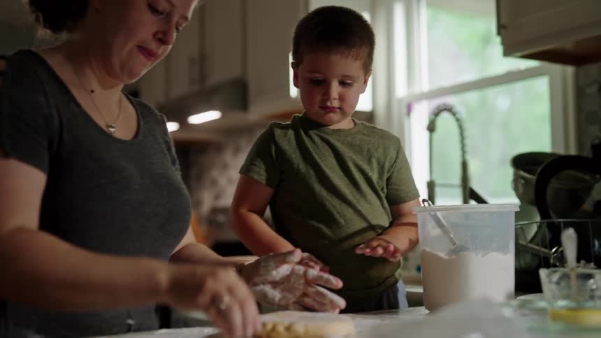 A mother and her young son are happily baking together in the kitchen with the mother kneading dough while the boy watches eagerly beside her