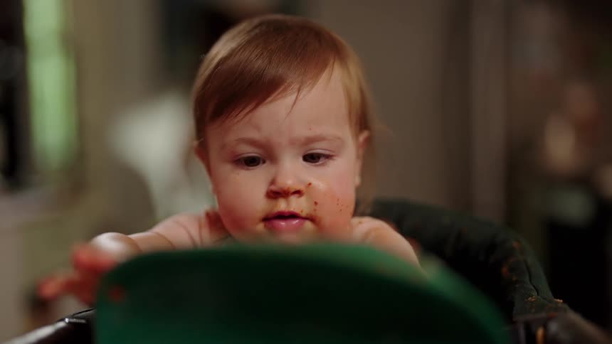 A baby with a messy face and a stained shirt is sitting in a high chair playing with a green plate that has a few spaghetti noodles on it picking it up and setting it back down