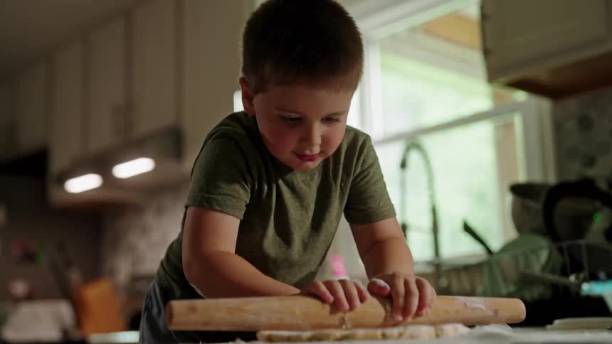 A mother has her son help out in the kitchen with the son rolling dough on the countertop using a rolling pin while the mother prepares another meal on the stove at midday
