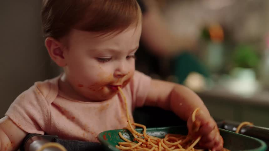 A small child in a high chair is messily eating a bowl of spaghetti with their hands pulling the noodles out of the bowl and putting them into their mouth as a parent stands in the background