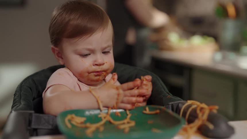 Toddler happily self feeding on a plate of noodles