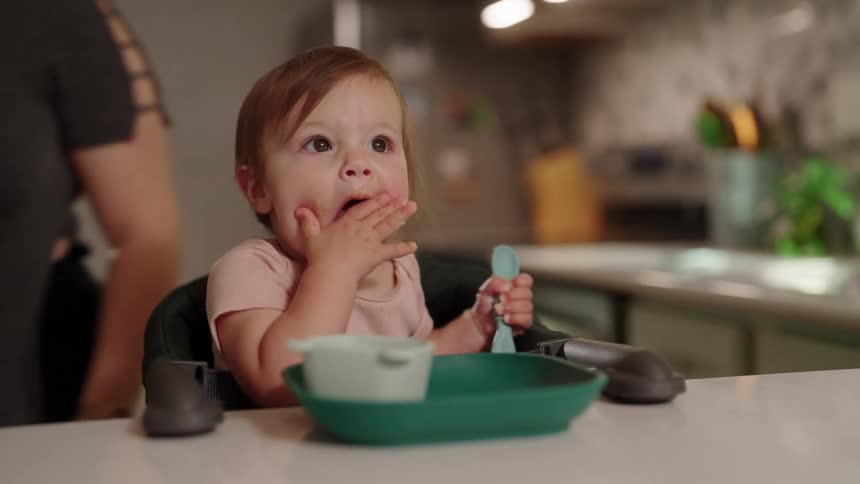 Baby sitting eating with her hands and her mother walking around the kitchen