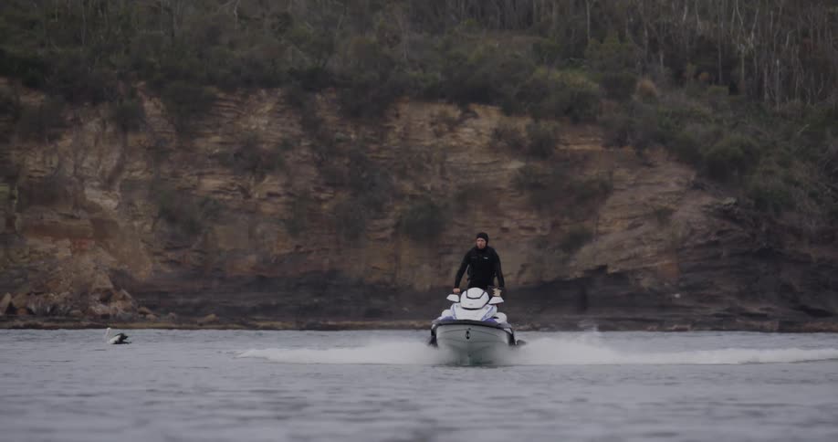 A man wearing a black swimming suit rides a jet ski in the ocean near the shore with ducks swimming in the water at midday