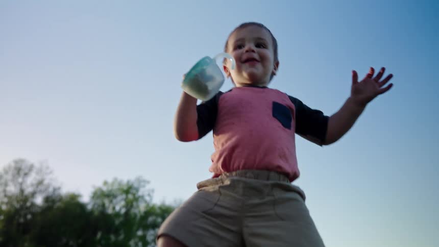 Toddler carrying a closed container gets up and falls more than once on the lawn in a park