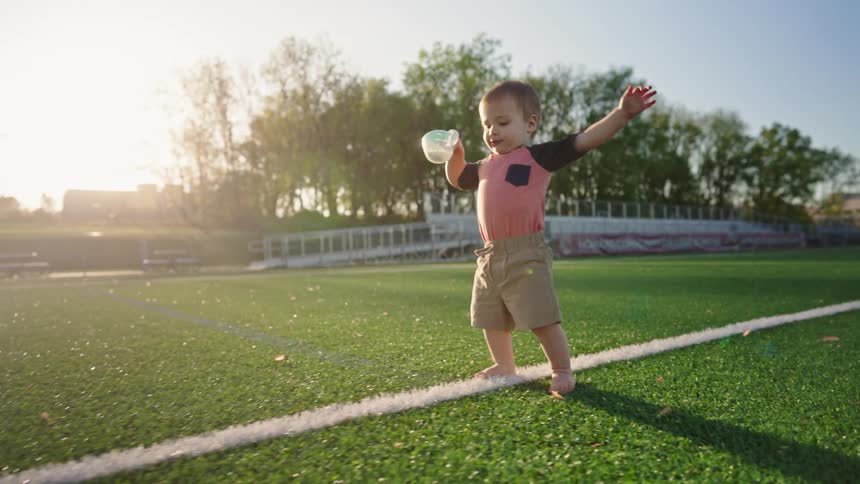 A barefoot toddler walks happily on a sports field holding a sippy cup and waving