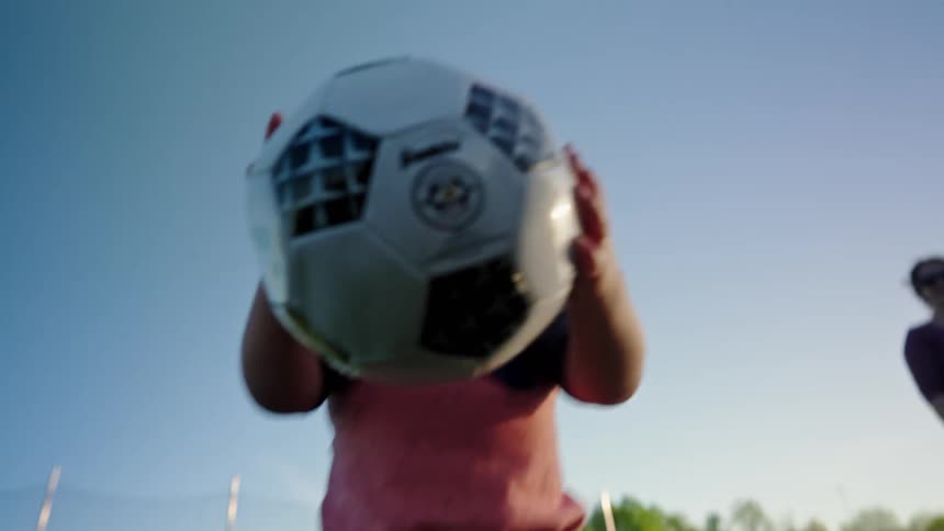 A small child in a red and black shirt and tan shorts is seen on a soccer field picking up a soccer ball holding it over their head and then dropping it back down to the ground