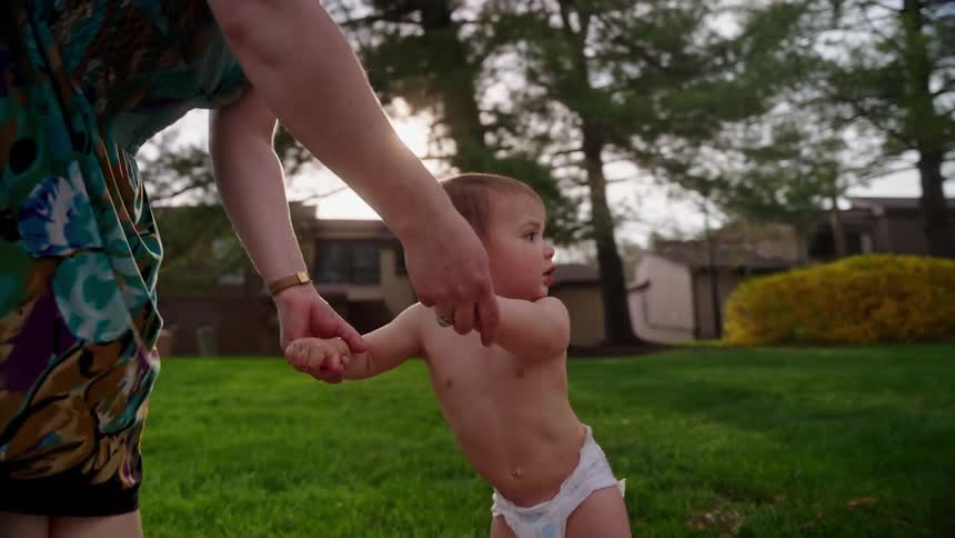A mother wearing a colorful dress is helping her baby who is wearing a diaper learn to walk on a sidewalk in a residential neighborhood on a sunny day