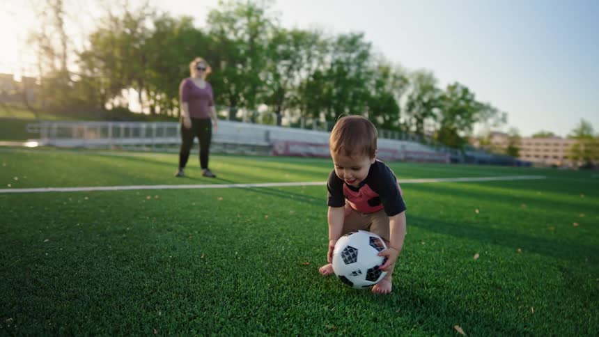 A playful toddler wearing a red and black shirt and tan shorts is seen on a sunny soccer field picking up a ball and dropping it as a parent stands nearby watching