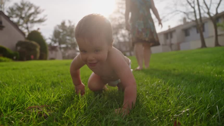 A baby in a diaper crawling on green grass outdoors with an adult standing in the softfocus background and the sun flaring brightly behind them
