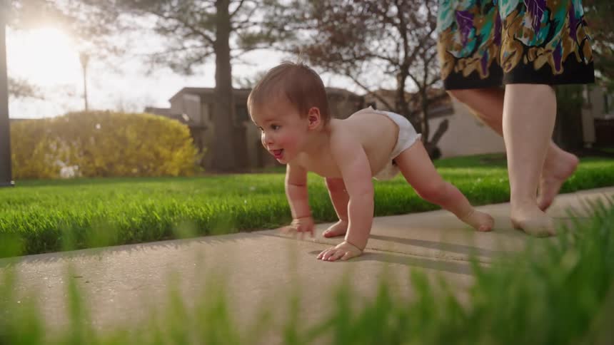 A baby crawls on a walkway while the mother walks behind him and picks him up to stand on his feet wearing only a diaper at midday