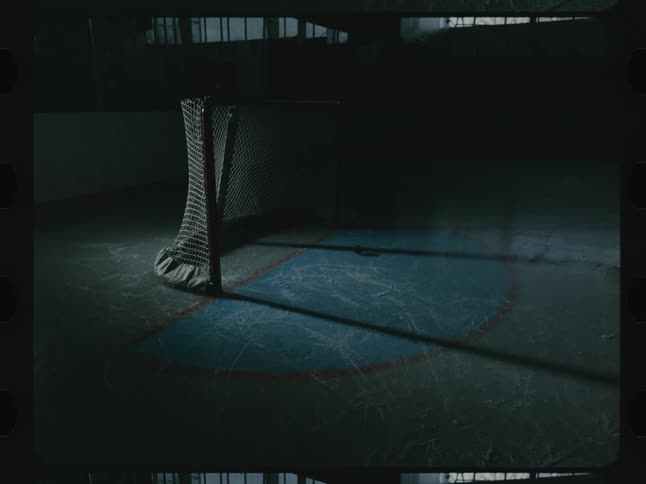 Hockey puck hits the goal net then rolls out across the ice of a shadowed indoor ice rink