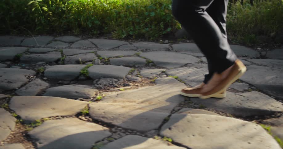 The video shows a lowangle shot of a persons legs and feet walking along an ancient stone pathway the camera follows the persons feet as they carefully step over the uneven stones and patches of grass