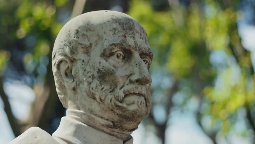 The video shows a closeup of a weatherworn white marble statue of a man with a beard and a somber expression the statue is surrounded by lush green foliage and sunlit trees which are slightly out of focus in the background