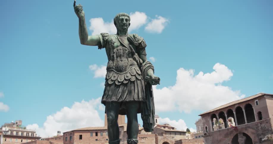 A large bronze statue of a roman emperor standing in a public square with ancient roman ruins and a clear cloudy blue sky in the background